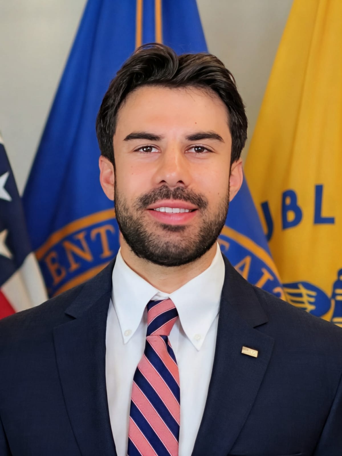 Noah Sofio, in a dark suit and striped tie, with flags behind him